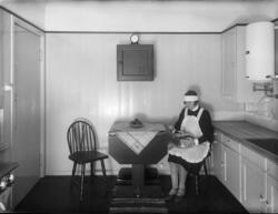 En kvinne ikledd hushjelpsuniform sitter i et ukjent kjøkken