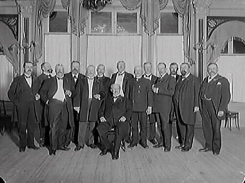 Gruppbild av uppklädda äldre herrar i sal med stiliga gardinuppsättningar.