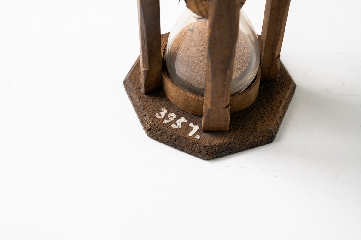 Timglas av glas, med sand som mätare, sitter i åttkantig träform som hålls ihop med fyra träribbor. Glaset hålls på plats med näver. Text på timglasets ovansida.