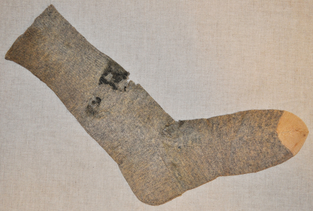En strumpa i ljusblått yllegarn med vit tå. Resårstickat ben och slätstickad fot. Fläckig och trasig.