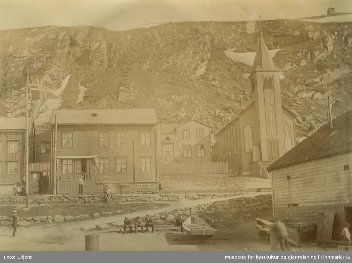 Den katolske kirken i Hammerfest i 1899. Til venstre for den står det trehus med skifertak. Foran husene står det en flagmast med et vaiende  norgesflagg. Nede i midten av bildet står en liten båt og ved siden av sitter tre men på en benk. Til venstre for dem er det en uniformert person. Arbeidsfolk er å se ved pakkhuset. Oppe på veien står det en kvinne og to barn. En man sitter med et barn i fanget på muren. Det virker som om innbyggerne i Hammerfest nyter godværet.  I fjellsida til Salen ligger det fortsatt litt sne. Oppe på Salenfjellet ser man varden.