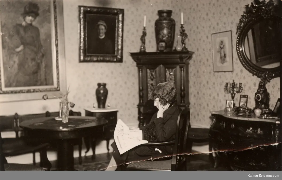 Fotografi av konstnärinnan Jenny Nyström i sin lägenhet. Hon är avbildad i profil med ansiktet vänt åt vänster. Hon sitter i en karmstol och läser en tidning. Framför henne står ett litet runt bord. Till höger om henne syns en annan karmstol. På väggen framför henne hänger självporträttet målat i Paris. I hörnan står ett hörnskåp. På skåpet står en urna och två ljusstakar. Till höger syns en rokokobyrå över den hänger en stor spegel med ram.