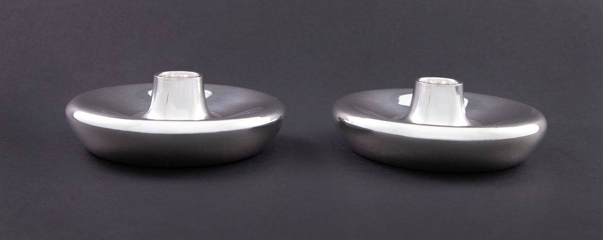 To lysestaker i sølv. Lav, sirkulært korpus med konvekst midtparti og sylindrisk lyspipe.