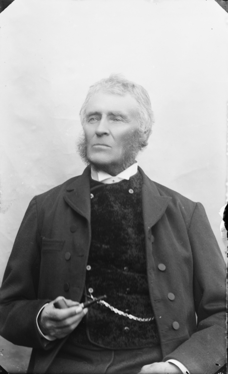 Mann i halvfigur med grått hår og stort kinnsjegg med pipe i handen, kledd i mørk vest og jakke