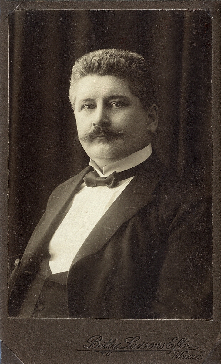 Porträttfoto av en medelålders man i mörk kostym med väst, stärkkrage och fluga.
Bröstbild, halvprofil. Ateljéfoto, 1912.