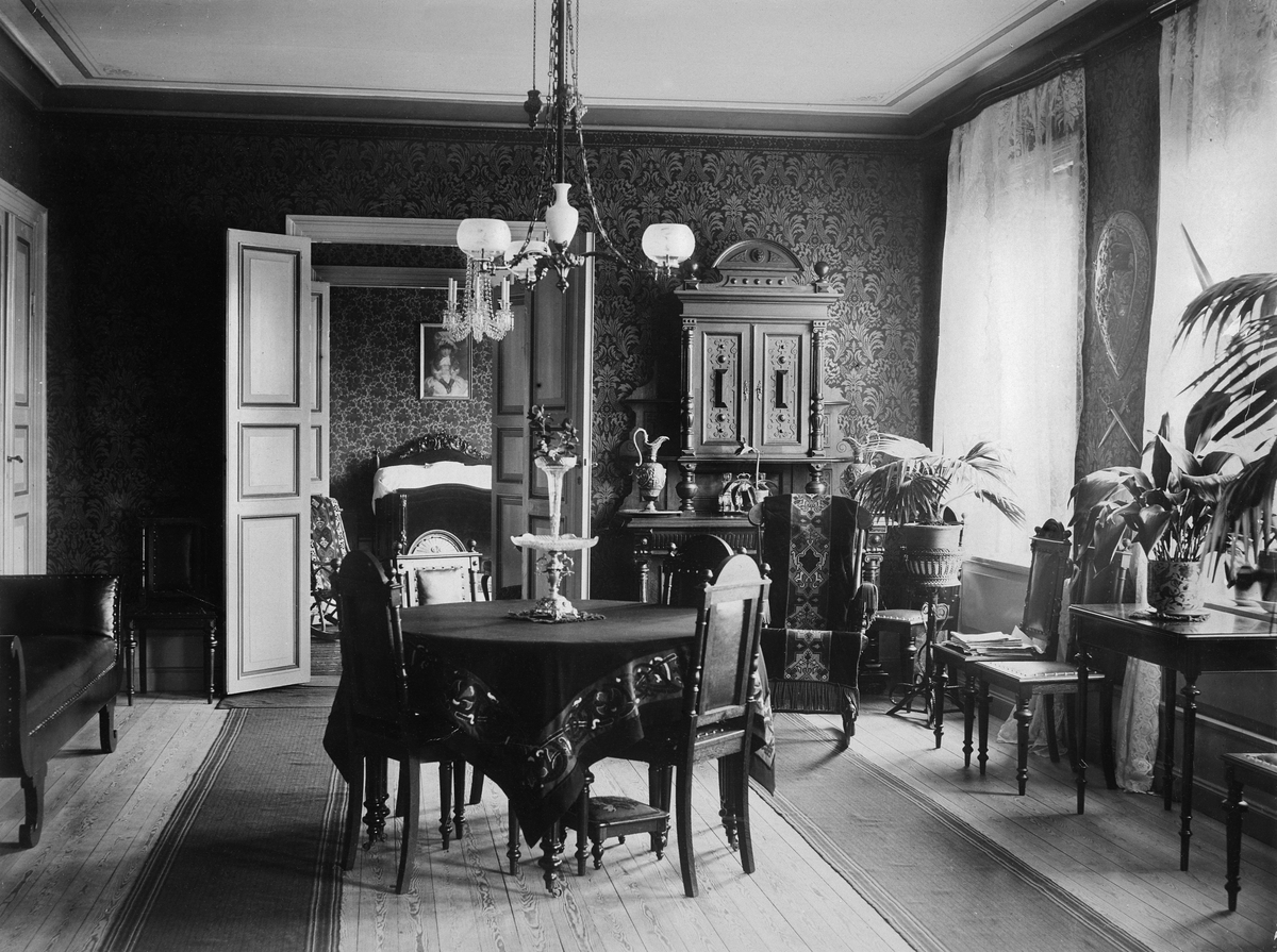 Interiör, borgerligt hem, ca 1890-tal. Matsal med ekmöbler i nyrenässans. Mörka tapeter, vita gardiner, på golvet endast gångmattor. Pinngungstolen är försedd med en broderad gungstolsmatta.
