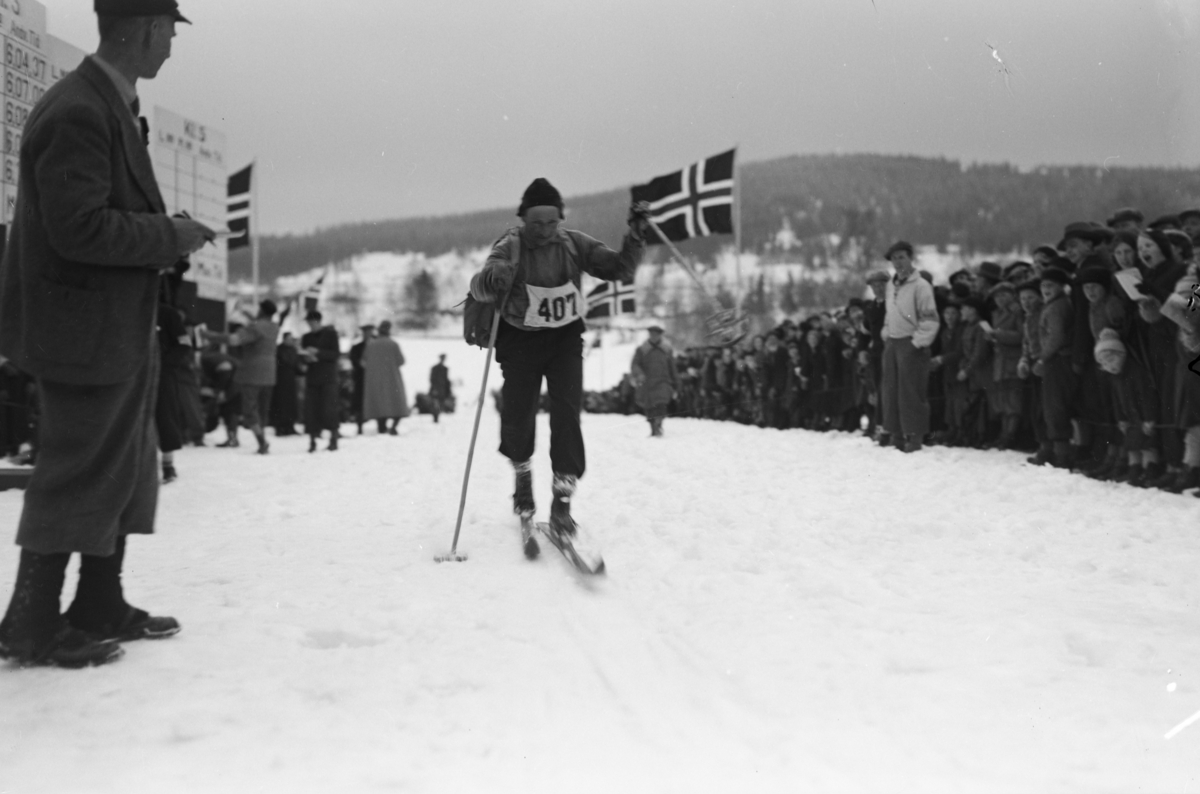 Birkebeinerrennet, start nummer 407 nærmer seg mål på Lysgårdsjordet i Lillehammer, tilskuere følger med. Mannen til venstre antas å være Lars Høgvold, formann i Lillehammer skiklubb. Rennet gikk fra Rena til Lillehammer.