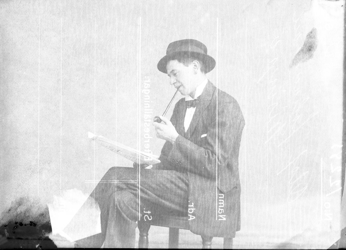 En ung man med hatt, pipa i munnen och tidning i knät sitter på en stol.