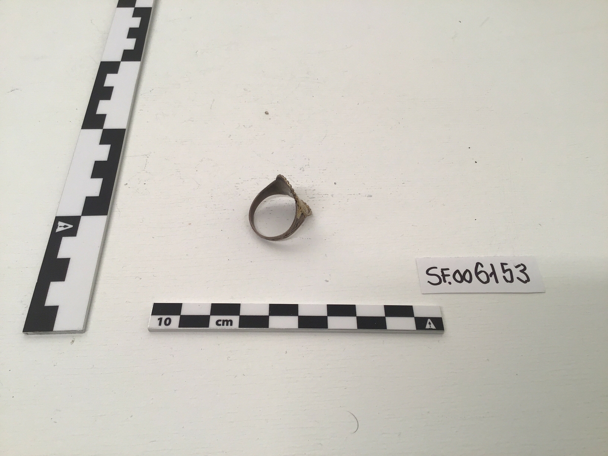 Fingerring av sølv med forgylt plate, plata er oval i foma med siselering. Plata går i ett med ringen og har enkel dekorkant.