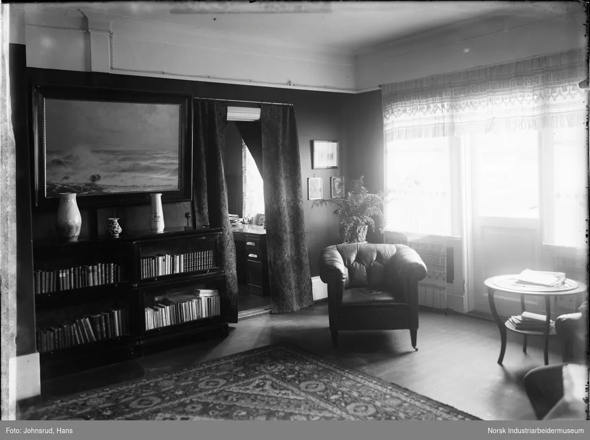 Interiør av stue. Midt i bildet står en stol med skinntrekk, Til venstre er det en veggseksjon med bøker under et maleri av havet. Mellom stolen og veggseksjonen er en dør til et annet rom. Til høyre er det et bord foran vinduer og en dør.