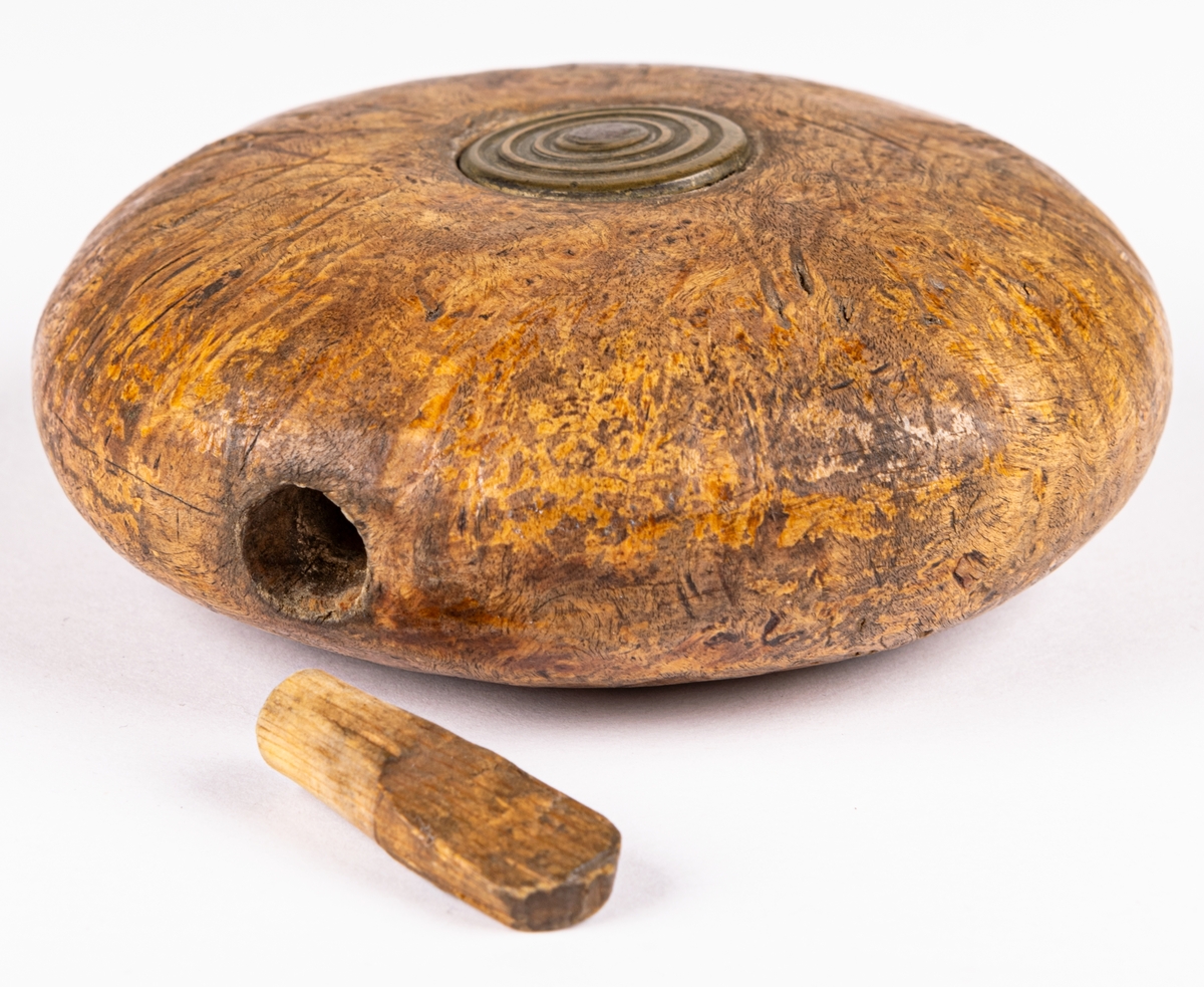 Snushorn, av masur, svarvat, rundat, platt, med ett runt mässingsbeslag på mitten av båda plattsidorna. I ena kanten hål för träplugg.