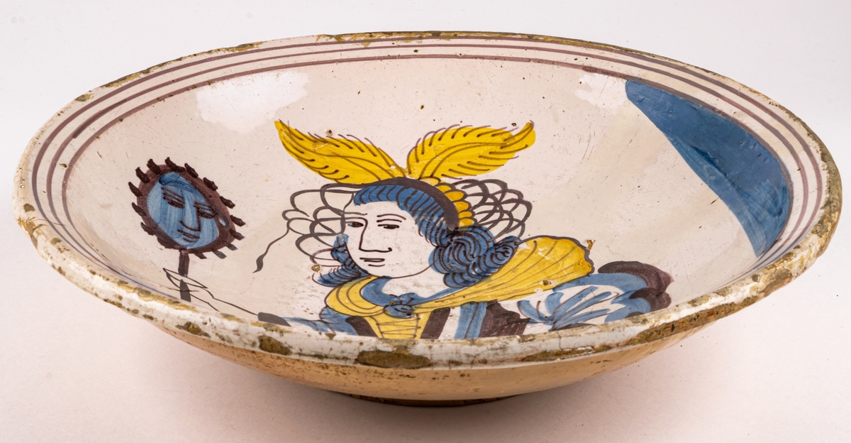 Keramikfat av ljusbrännande lergods med ljus tennglasyr som bottebfärg. Handdekorerad med kvinnogfigur som håller en spegel i högra handen. Kvinnofiguren har vingar på huvudet, snörliv och vid kjol. Figuren är i färgerna brun-lila, gult och blått.
Fatet är troligen från 1700-talet.
En spricka i fatet som löper genom en tredjedel, små naggar i kanten samt