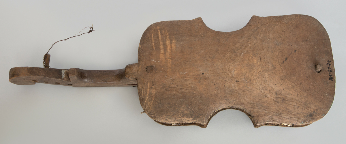 Fragment. 4-strenget. Laget av ubehandlet tre, skåret med kniv. Meget primitivt. Form omtrent som en vanlig fiolin, men svært lite hvlevet bunn og lokk.
