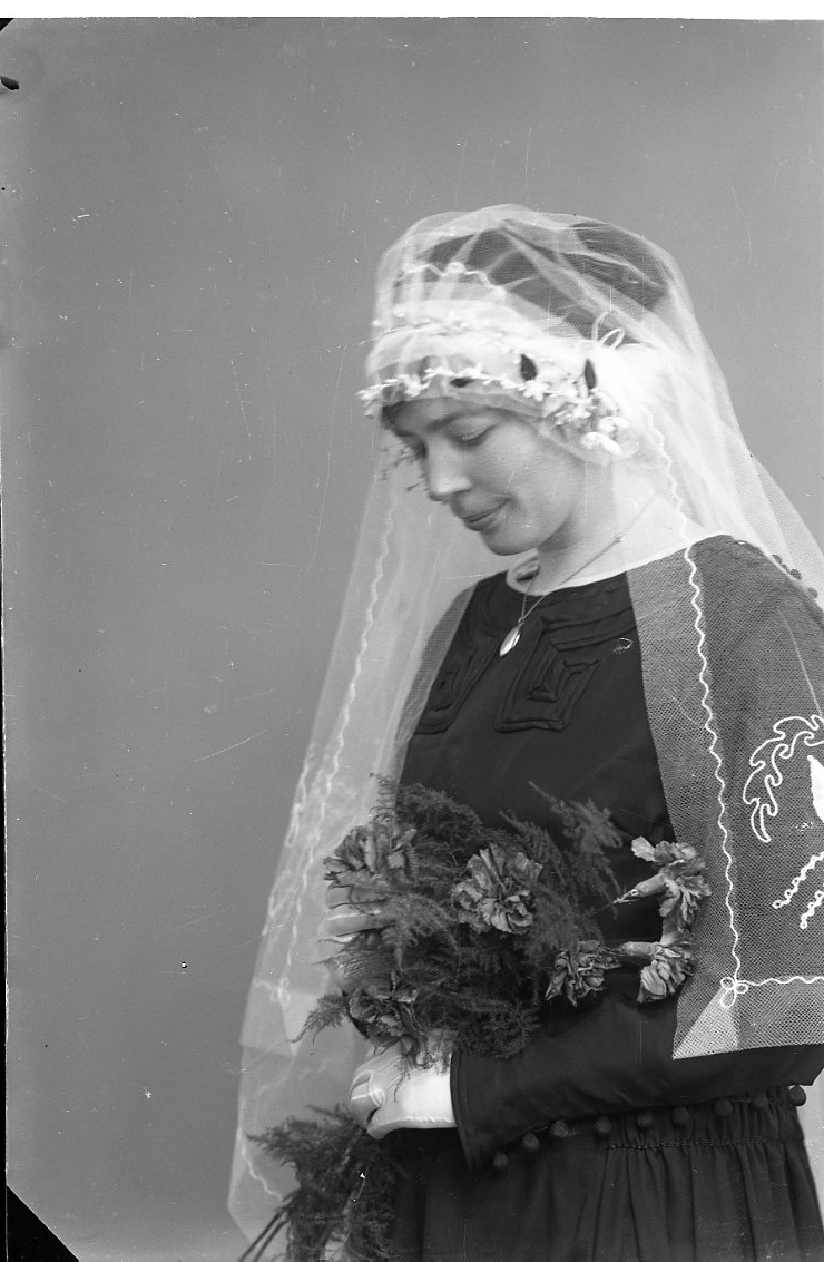 Porträtt av en brud i 3/4-profil. Hon bär mörk klänning, flor och handskar. Hon tittar ner mot en brudbukett hon håller i händerna.