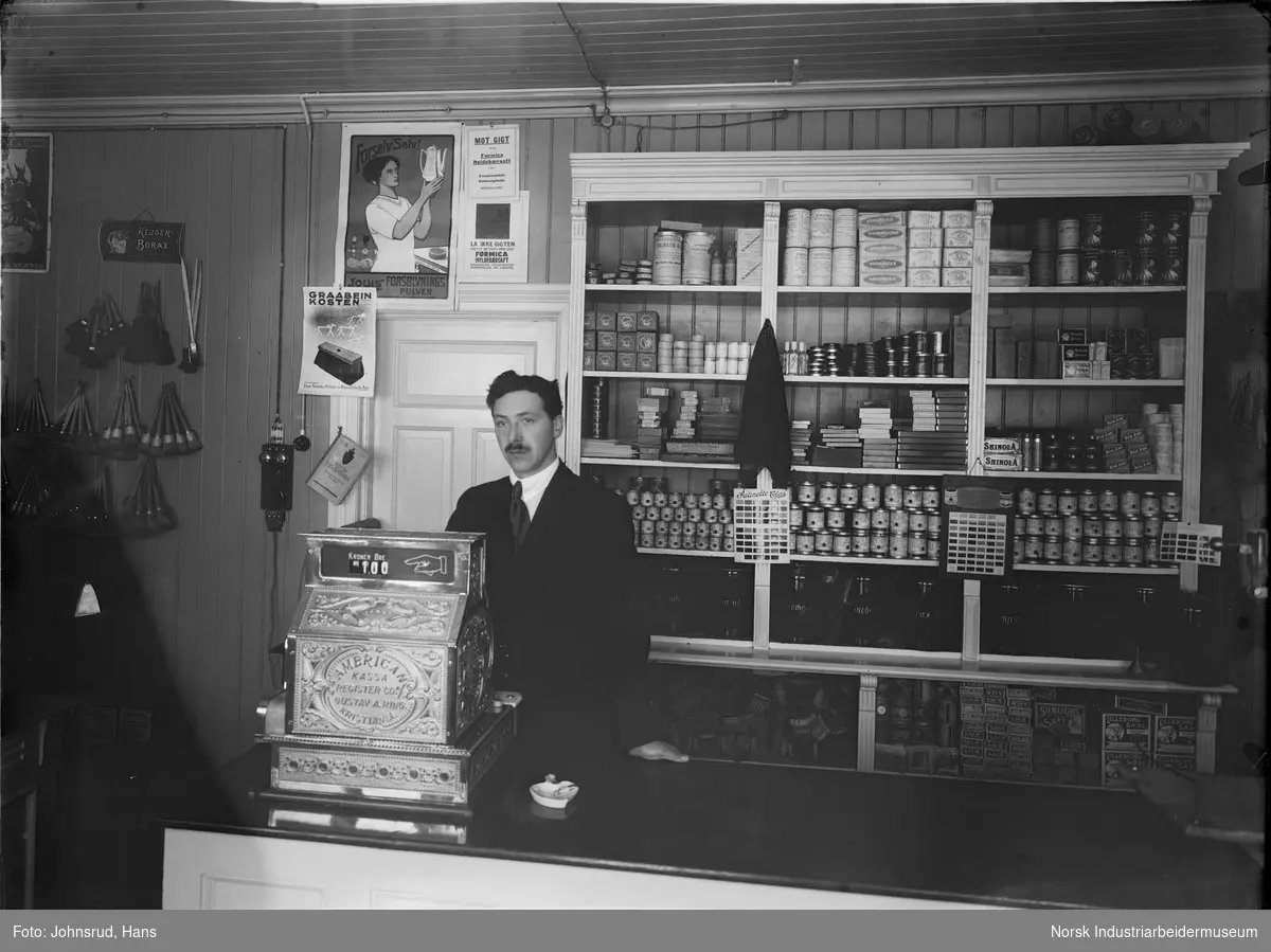 Interiør av butikk. En mann står med kassa bak disken. Hyllene på veggen er fylt med forskjellige varer. Til venstre i bildet henger det malingskoster på veggen.
