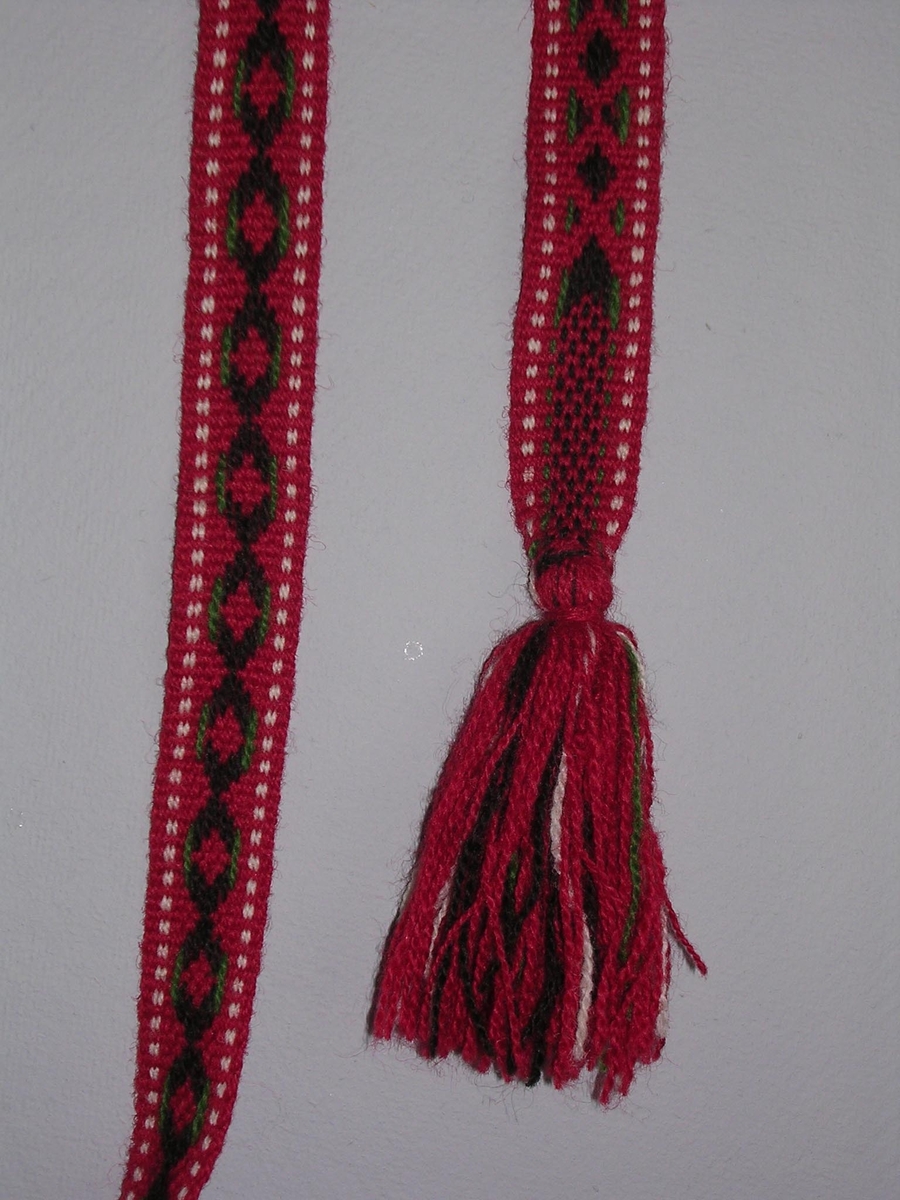 Mönstervävt band, handvävt, i rött, svart, vitt och grönt ullgarn. Tofsar i båda ändar, ca 10 cm långa.

Funktion: Järvsö, Hälsingland