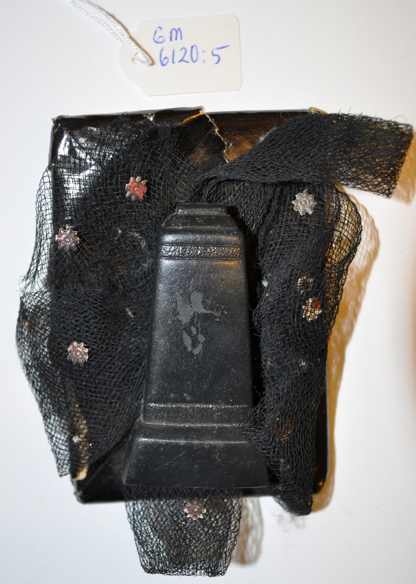 Rektangulär begravningskonfekt inslagen i svart glanspapper. På en svart tyllrosett dekorerad med små pappersblommor finns en gravsten i svart gjutet material. Pappret är trasigt och man kan se själva karamellen.