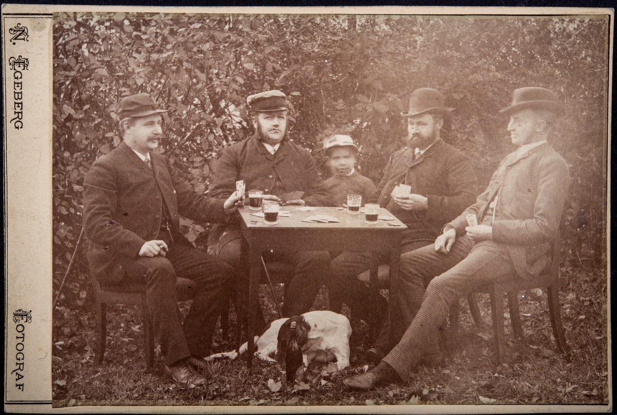 Gruppe 4 menn som spiller kort, kortspill. Nr 2 fra venstre er Andreas Smestad (1845-1892) fra Smestad Østre på Ring i Ringsaker. Et barn står å ser på, en hund ligger under bordet.