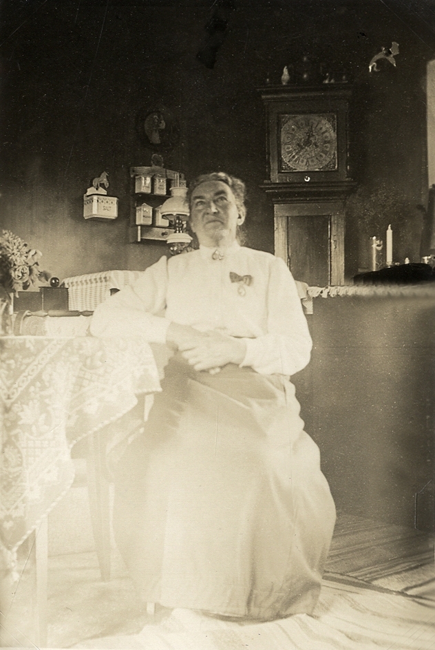 En medelålders kvinna, Lovisa Kullberg ("Lovisa te Boismans"), med förtjänstmedalj på bröstet, sitter vid ett köksbord, troligen i Boismanska gården, Växjö, ca 1915.
