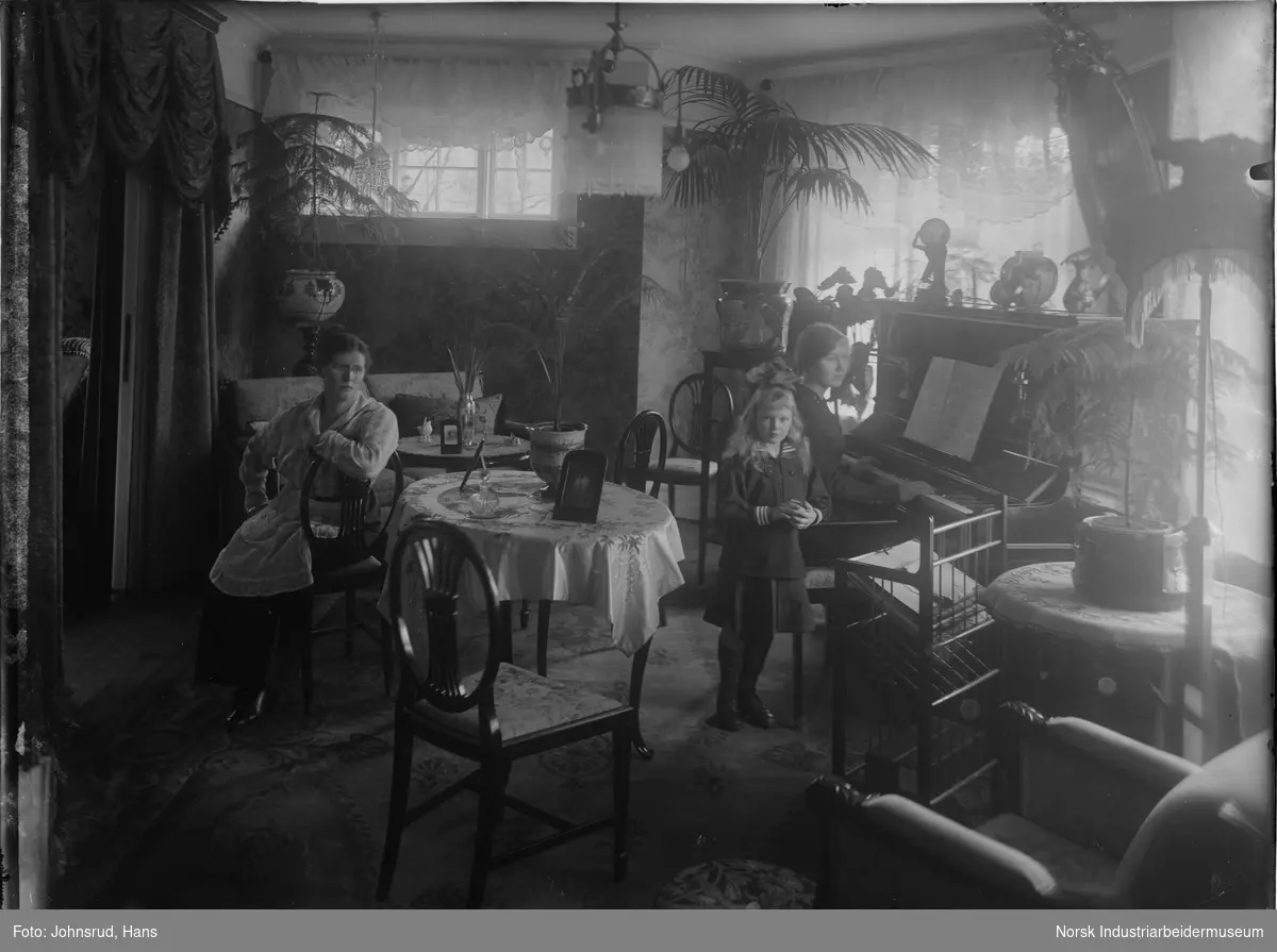 To sittende kvinner og en stående jente i en stue. Den ene kvinnen sitter ved et bord midt i bildet. Den andre kvinne sitter ved et piano i høyresiden av bildet. En sofagruppe er synlig i bakgrunnen. En lenestol er synlig i forgrunnen.