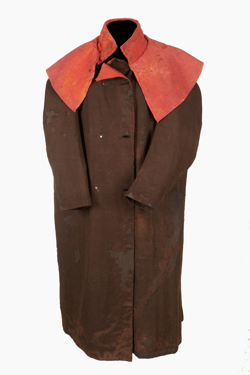 Kyrkvaktarrock av brunt grovt kläde med röd krage och ärmuppslag av vadmal. Tre knapphål sydda med svart tråd. Sprund bak.