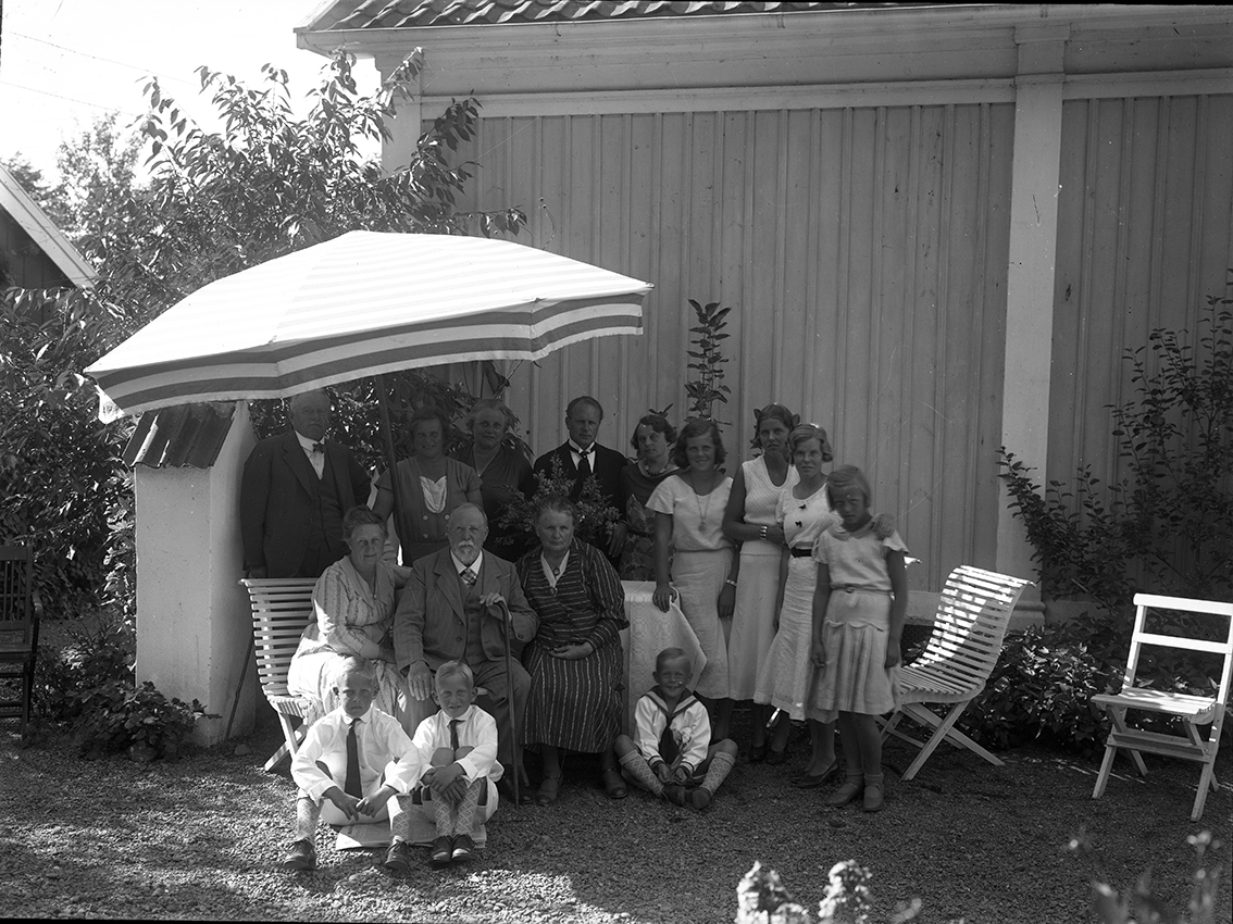 Ett sällskap med vuxna och barn sitter i skuggan av ett parasoll utmed en husvägg. Det är herr och fru Oldenburg med barn och barnbarn.