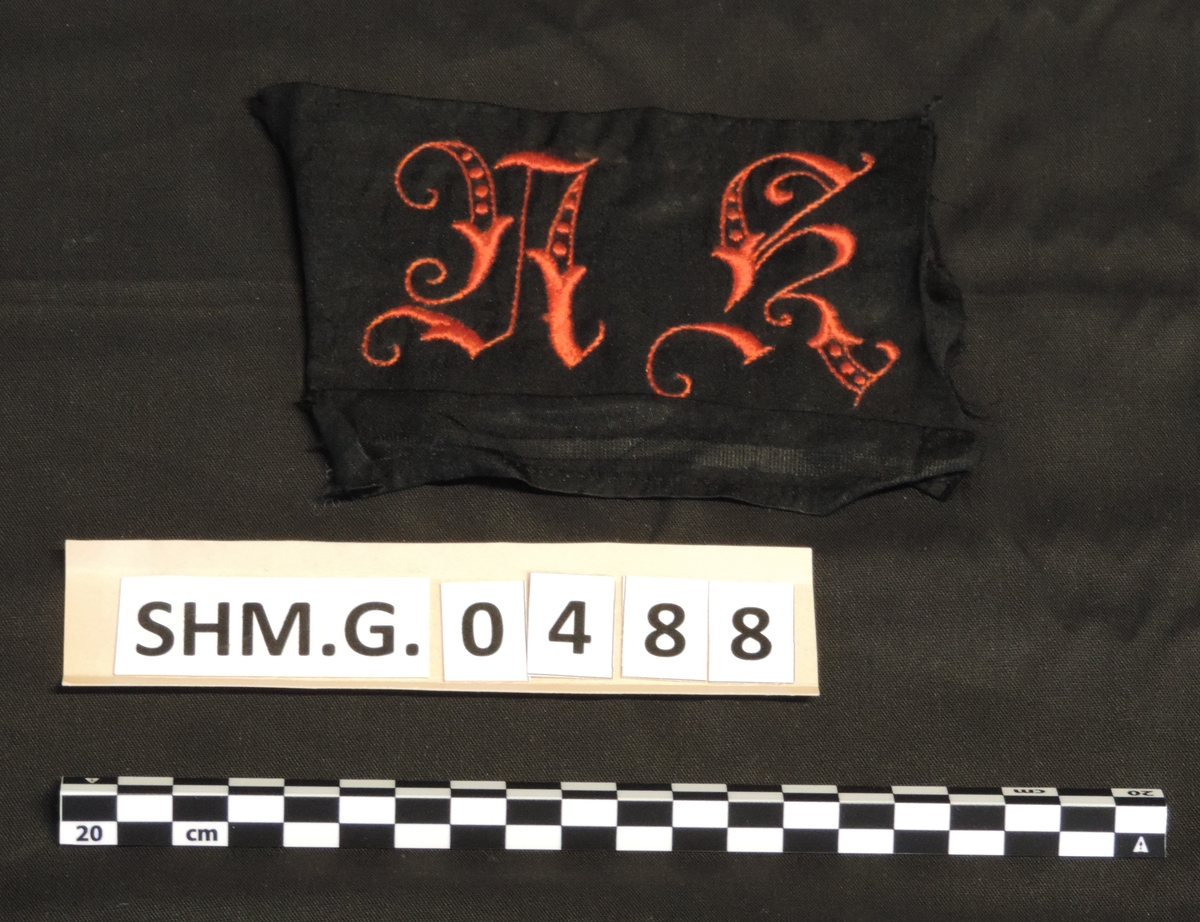 Kåpeskjold i sort silke, initaialer brodert i rødt AK