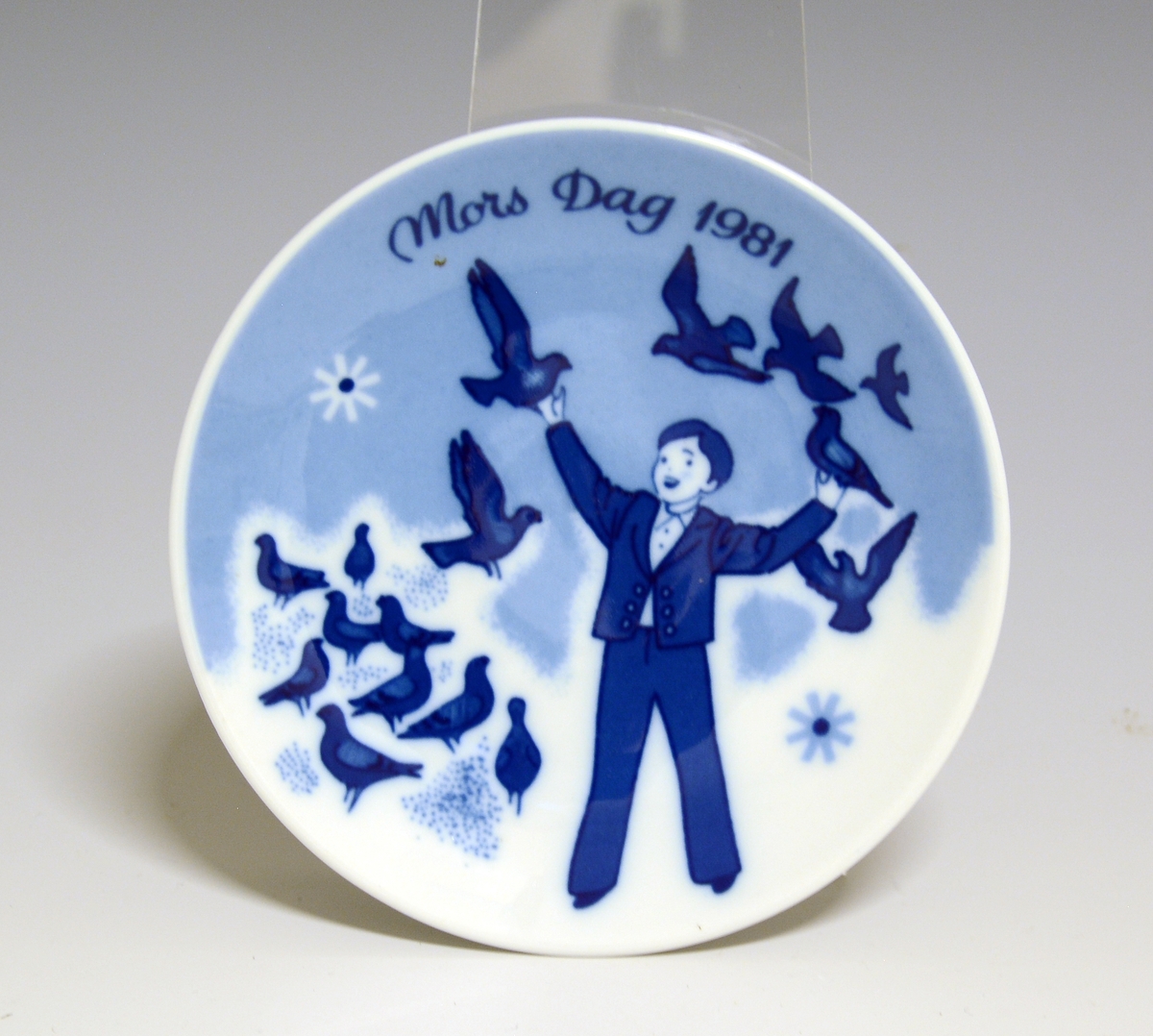 Morsdagsplatte av porselen. Hull til oppheng bak. Hvit glasur. Dekorert i blått med en gutt omgitt av duer, tekst Mors Dag 1981.