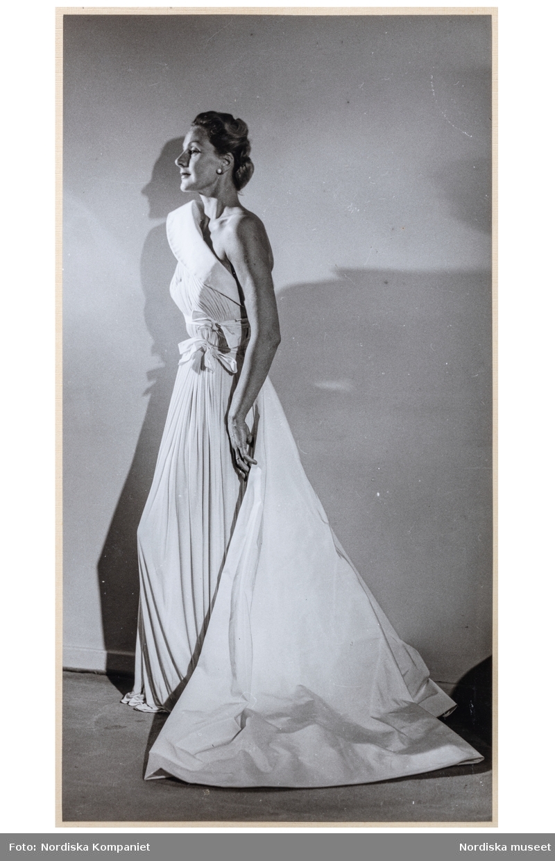 Mannekängen Margareta Berglund gift Nordlund (1946) visar festplagg i provrummet vid NK:s Franska damskrädderi. aftonklänning med ena axeln bar, nedvikt krage, rosetter vid midjan. Plisserat tyg, hellång kjol. Släp av slätt tyg.