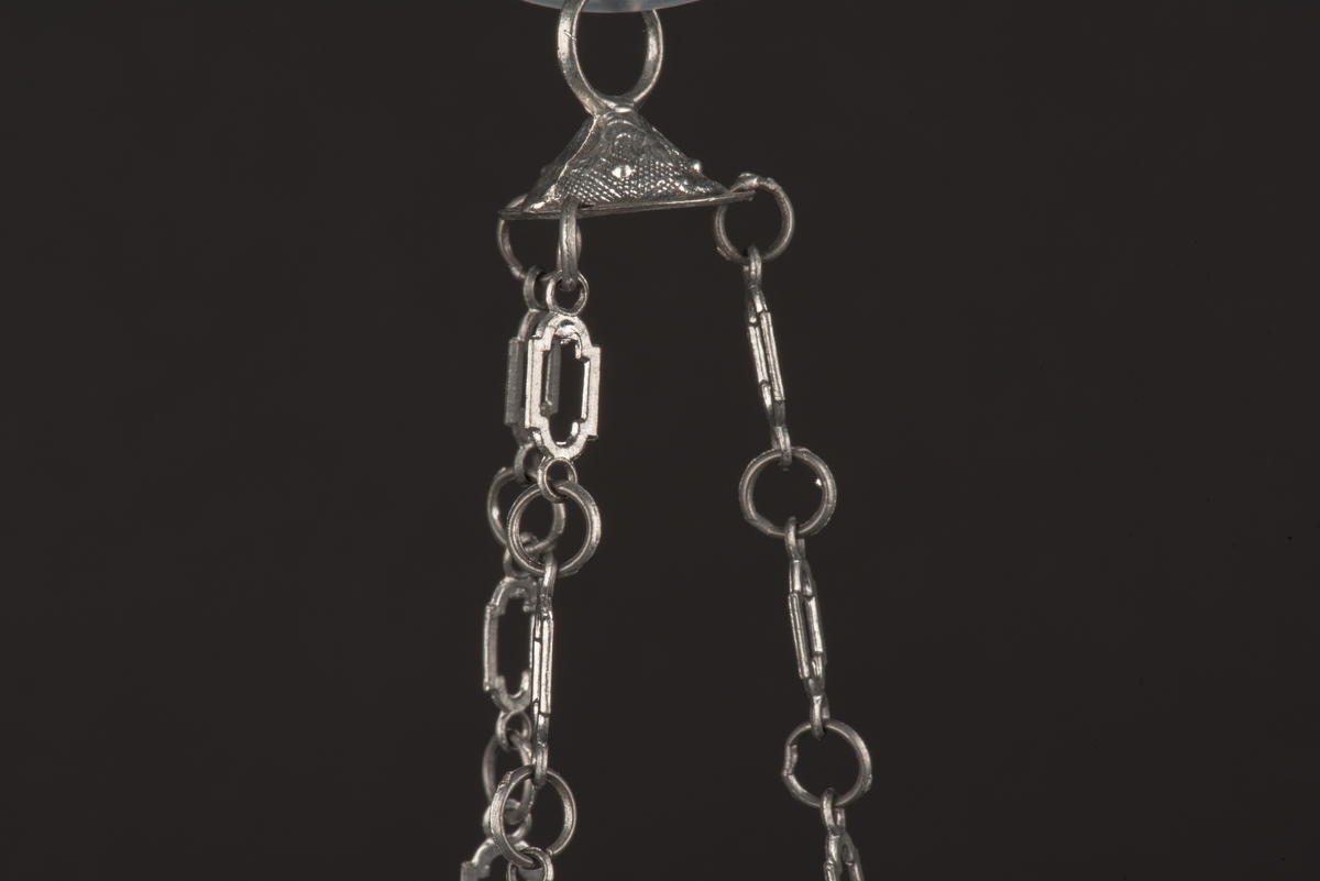 Ljuskrona till dockskåp, tillverkad av tenn.
Kronan har 6 ljusarmar, något tillböjda, och hänger i 3 kedjor.