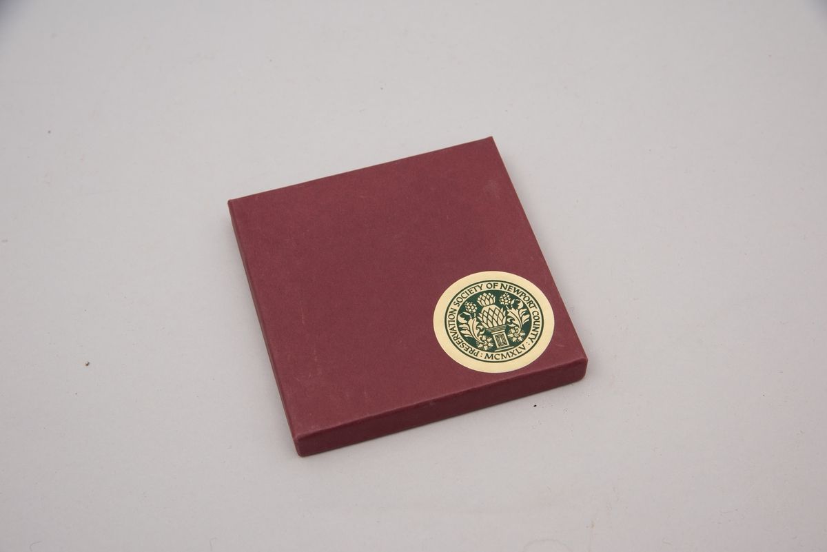 Marmorflis og souvenir fra Newsport Mansions med påført slagord "Well Behaved Women Rarely Make History". Tilhørende eske og informasjonsfolder. Mottoet påført marmorflisa er i ettertida blitt brukt til å beskrive Alva Vanderbilt Belmont (1853-1933) og andre kvinner som kjempet for kvinners stemmerett på begynnelsen av 1900-tallet i USA under slagordet "Votes for Women". Selve "Votes for Women"-mottoet ble brukt i 1909 på et berømt "open house" stemmerettsmøte holdt på Marble House, Newport, Rhode Island".