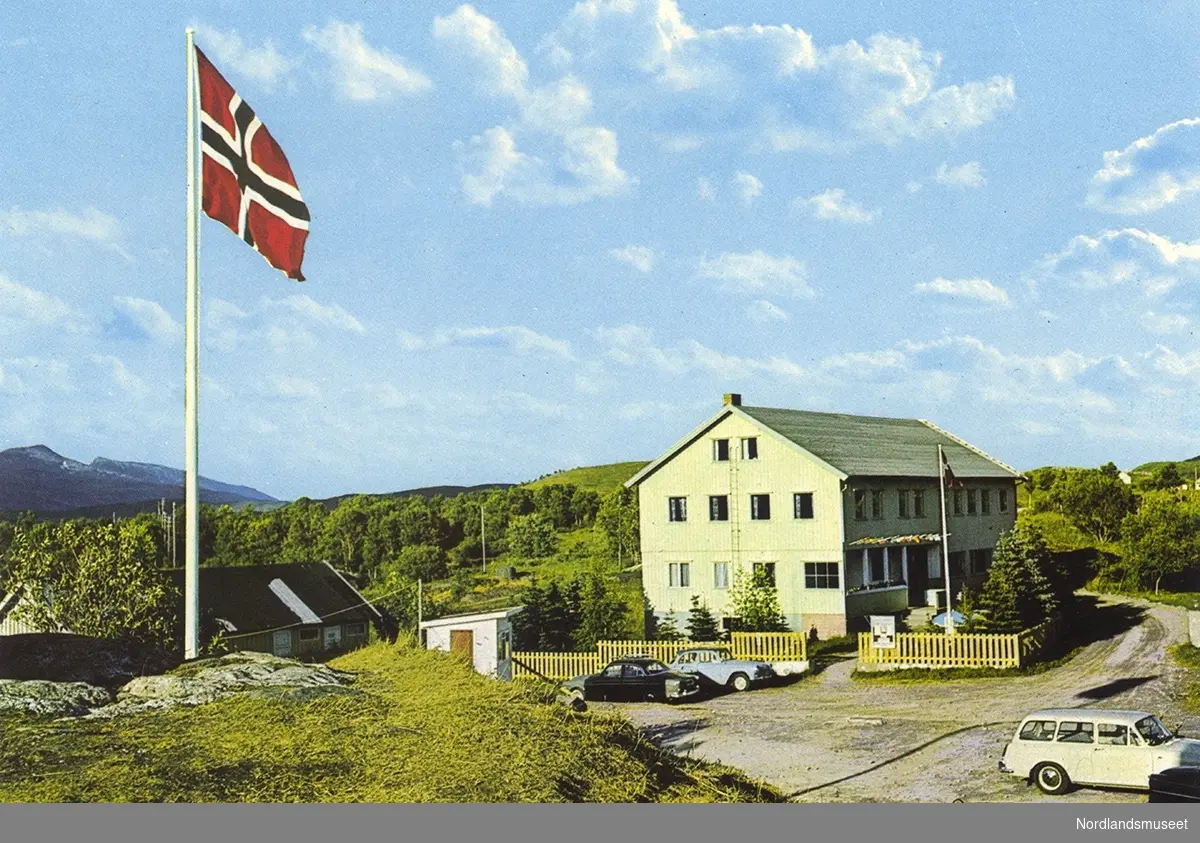 Fra Saltstraumen turisthotell. Større bygning. Hagegjerde. Parkeringsplass og noen biler. Bygning til venstre. Trær og fjell i bakgrunnen. I forgrunnen flaggstang med det norske flagg heist.