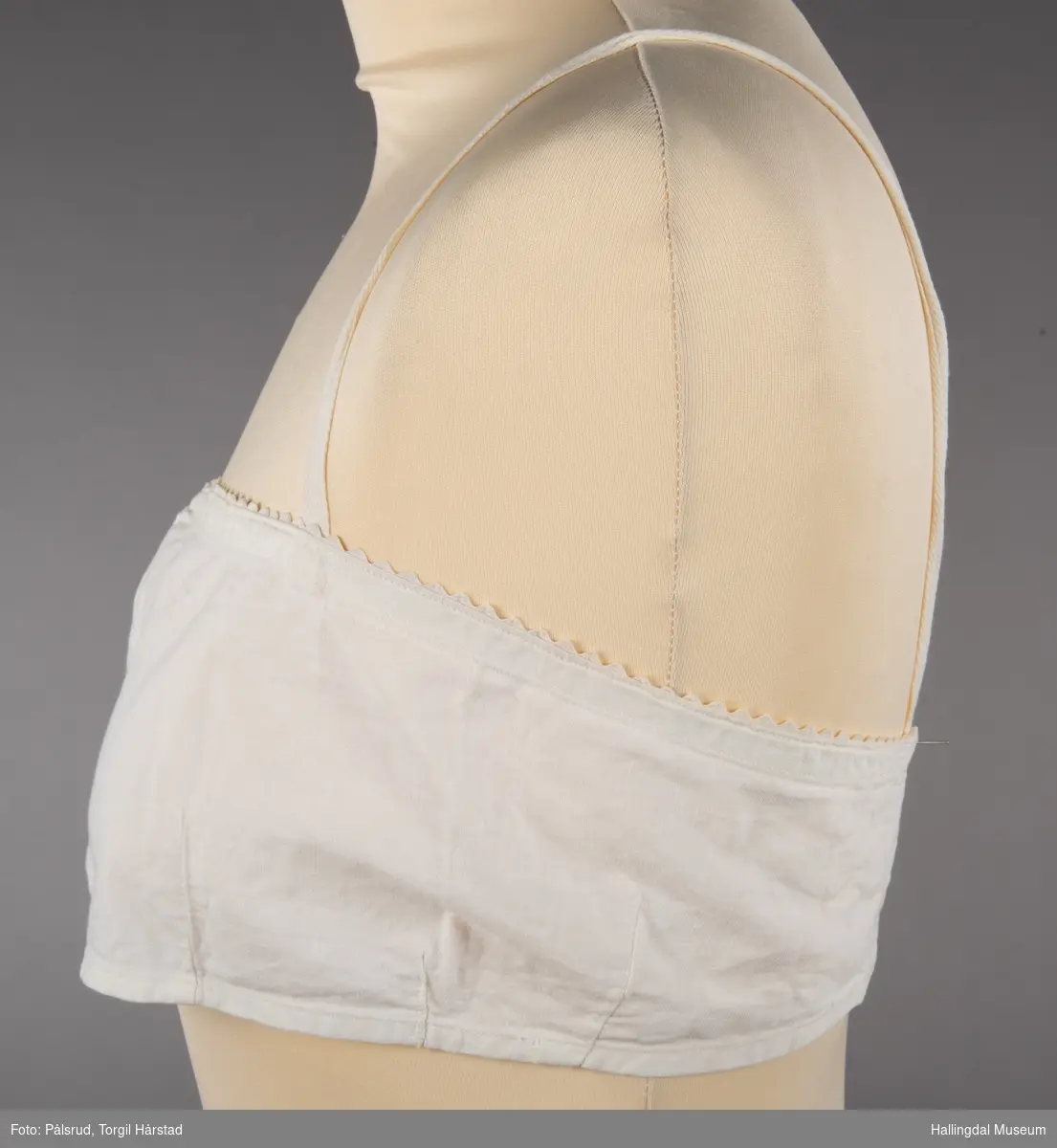 En hvit brystholder i bomull med hvit kroklisse i øvre linning. To enkle skulderstropper. To hvite perlemorsknapper bak som festes i enten to hemper eller to knapphull.