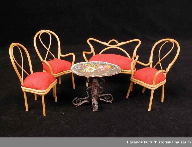 Dockskåpsmöbler i trä och röd textil. Stolarna och soffan är stoppad med halm/träull.
a) soffa. b 1-3) stolar. c) bord med pappskiva smyckat med blommor.