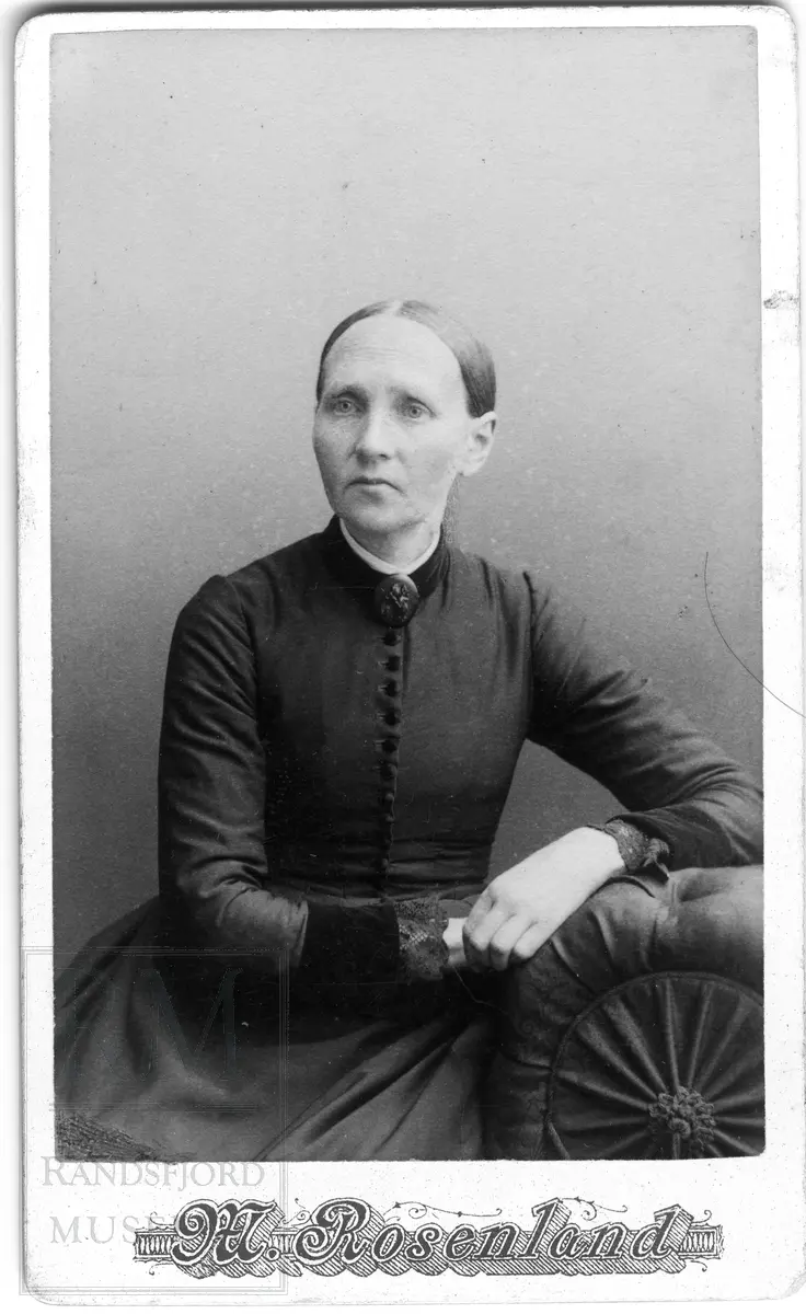 En kvinne i mørk kjole med knapper i front sitter og lener seg mot ei stoppet pute/lene. Stor brosje i halsen.