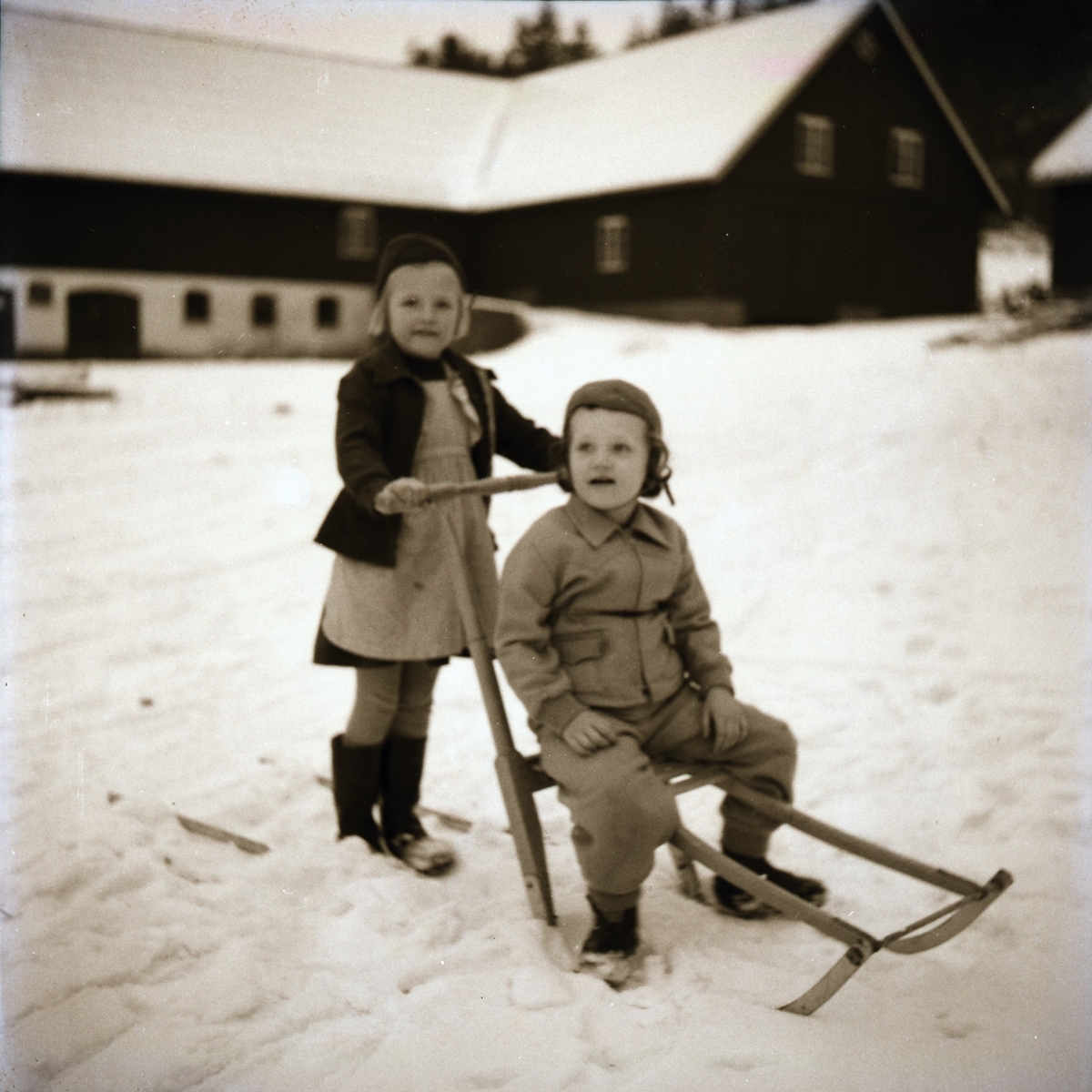 Vinterbild från Kantaburs gård i Veddige. Flickan Elin står med en sparkstötting, där troligen hennes bror sitter. I bakgrunden en stor ladugård. (Se även bild EA0197)