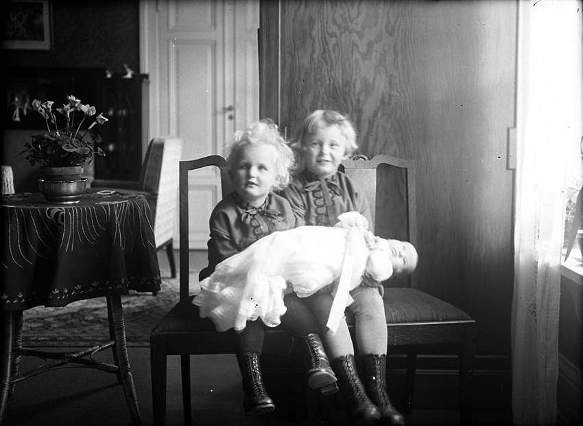 Två småflickor sitter på varsin stol med en baby, klädd i dopklänning,  liggande över knäna. Västerås.