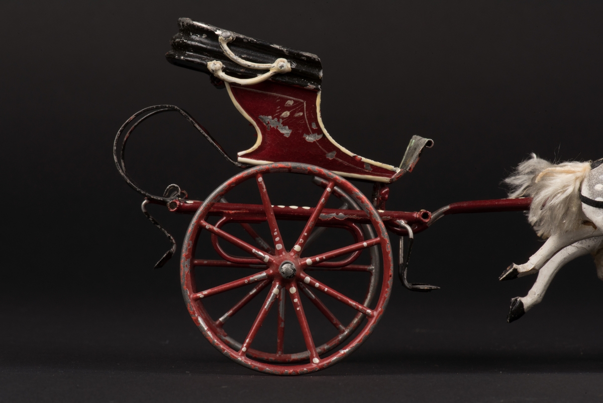Leksaksekipage som består av en vagn av plåt, dragen av två hästar av trä.
Vagnen är tillverkad av plåt och målad i rött, grönt och svart. Vagnen är öppen med nedfälld sufflett, ensitsig och med två hjul.
På sittplatsen finns en nedböjd metallten där den ursprungliga dockan sattes.
Vagnen är förspänd med två apelkastade hästar, vita och grå med fläckar. Hästarna är av trä överdragna med en massa. Man och svansar är av silke, seldonen av läder.
Mellan hästarna finns en hjuldriven funktion som gör att hästarna rör sig upp och ned och ser ut att trava när man skjuter ekipaget framåt.