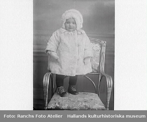 Ateljéporträtt av ett vinterklätt barn som står i en stol. Barnets mor är Fru Nilsson, Trikåfabriken i Varberg.