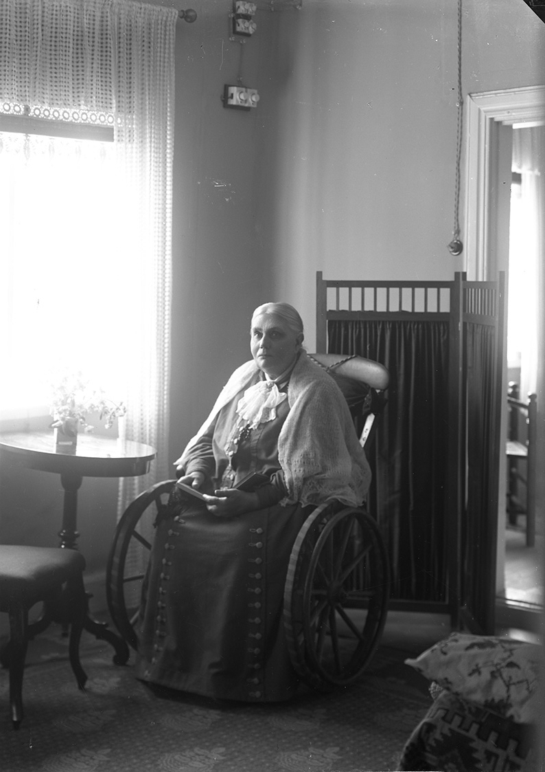 Porträtt av en kvinna som sitter i en rullstol med uppslagen bok i knät. Hon har en virkad sjal över axlarna.