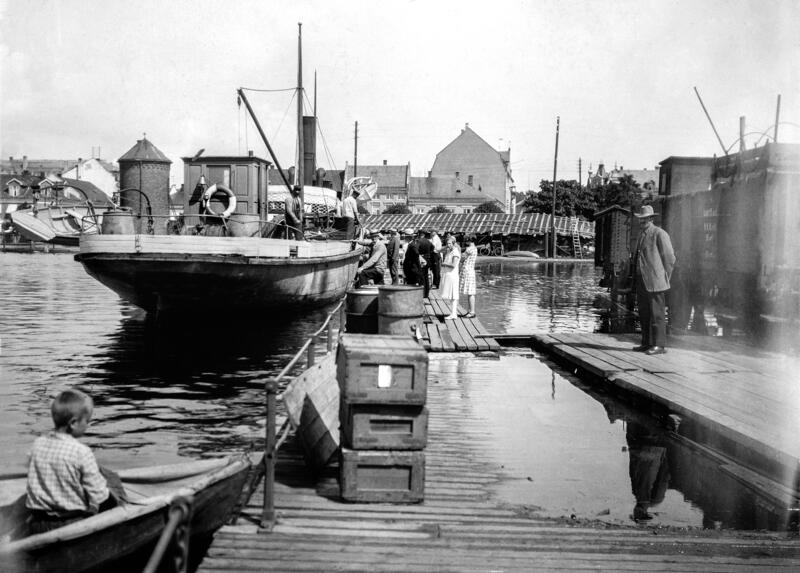Fotografi av en båt med stavnen mot fotografen, personer som går ombord og en jernbanevogn til høyre i bildet. Strandgata i Hamar skimtes i bakgrunnen. (Foto/Photo)