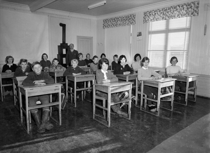 Svart-hvitt foto av et klasserom hvor elevene sitter på rekke bak pultene sine, bakerst i klasserommet står en etasjeovn.