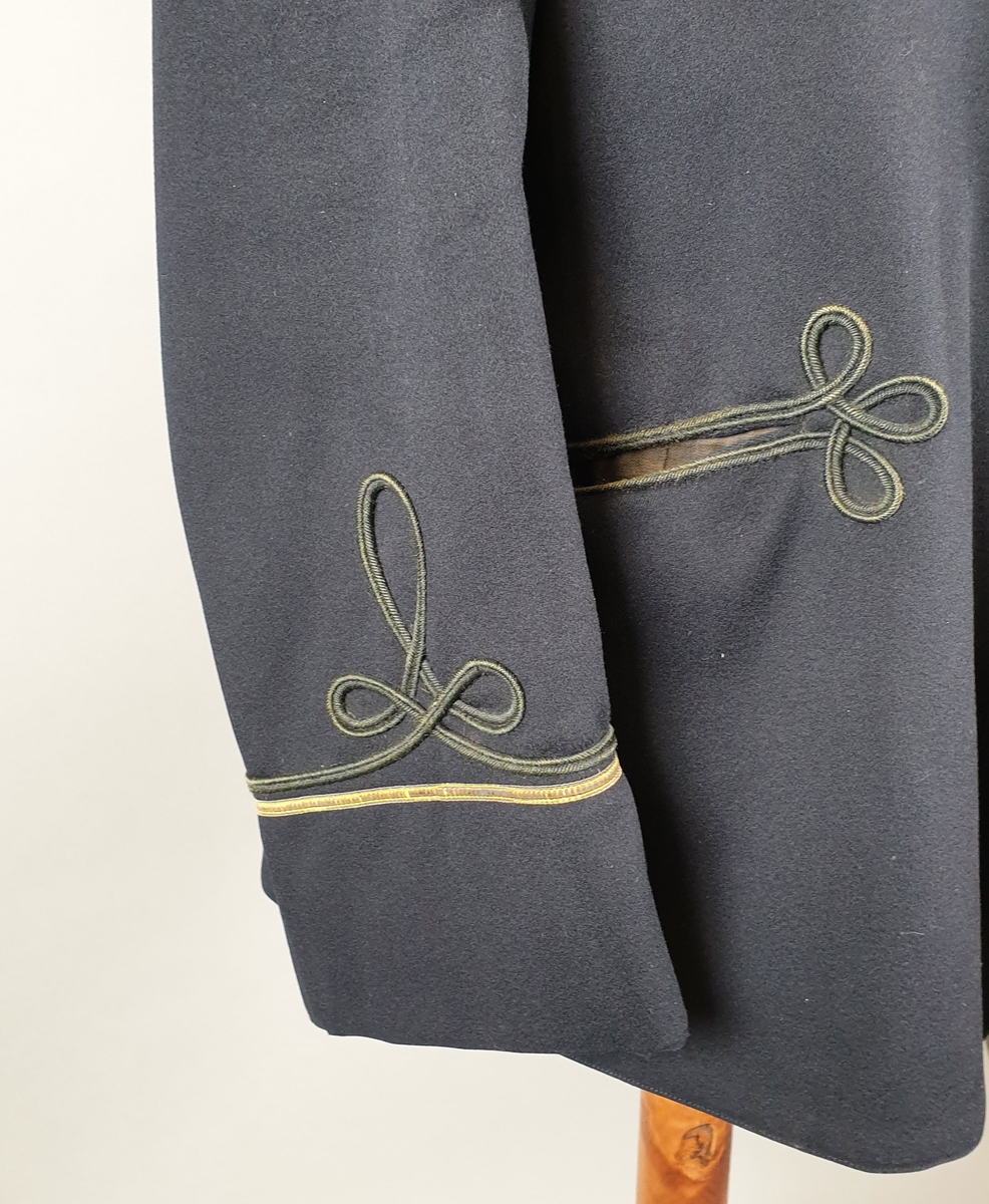 Svart uniformsjakke av ull, med skjulte knapper i front og grønt lissebånd som dekor på innsydde lommer på begge frontstykker og nederst på hvert erme. Gullbånd på hvert erme. Jakken er fôret med svart silke.