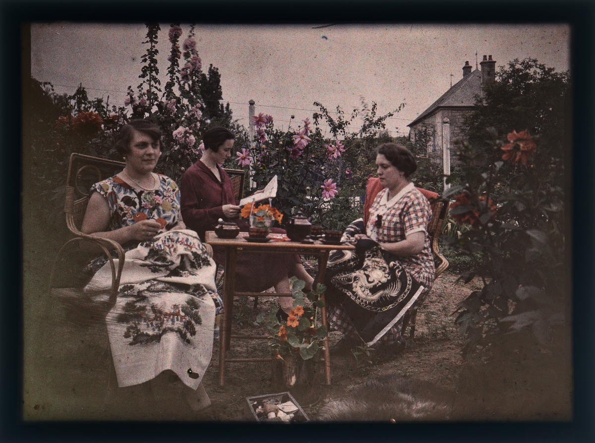 Tre kvinner som sitter ved et bord i en hage. To av dem syr/broderer og den andre leser.