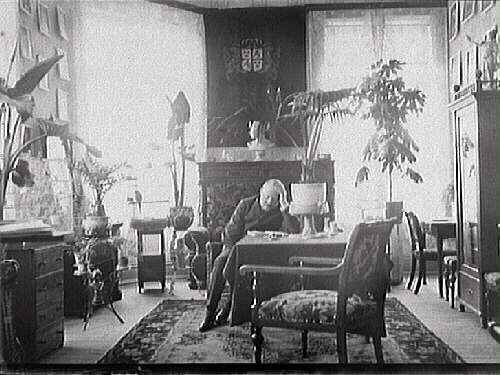 Interiör av en salong där en man sitter med huvudet lutat i ena handen vid ett bord. Rummet har mycket stora fönster, tidstypiska  gröna växter och många oramade bilder på väggarna.