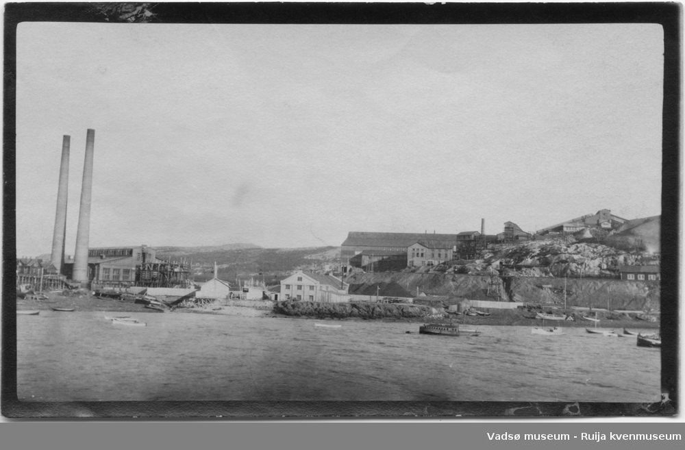 Bilde tatt fra sjøsiden, av Kirkenes havn. Flere av Sydvarangers  anleggsbygninger, og fabrikkpiper kan ses. Det ligger flere trebåter ute på sjøen. Bildet har tilhørt Lars Rygh fra Hønefoss. Han var med på mineryddingsoppdrag i Varangerfjorden 1917 - 1918.