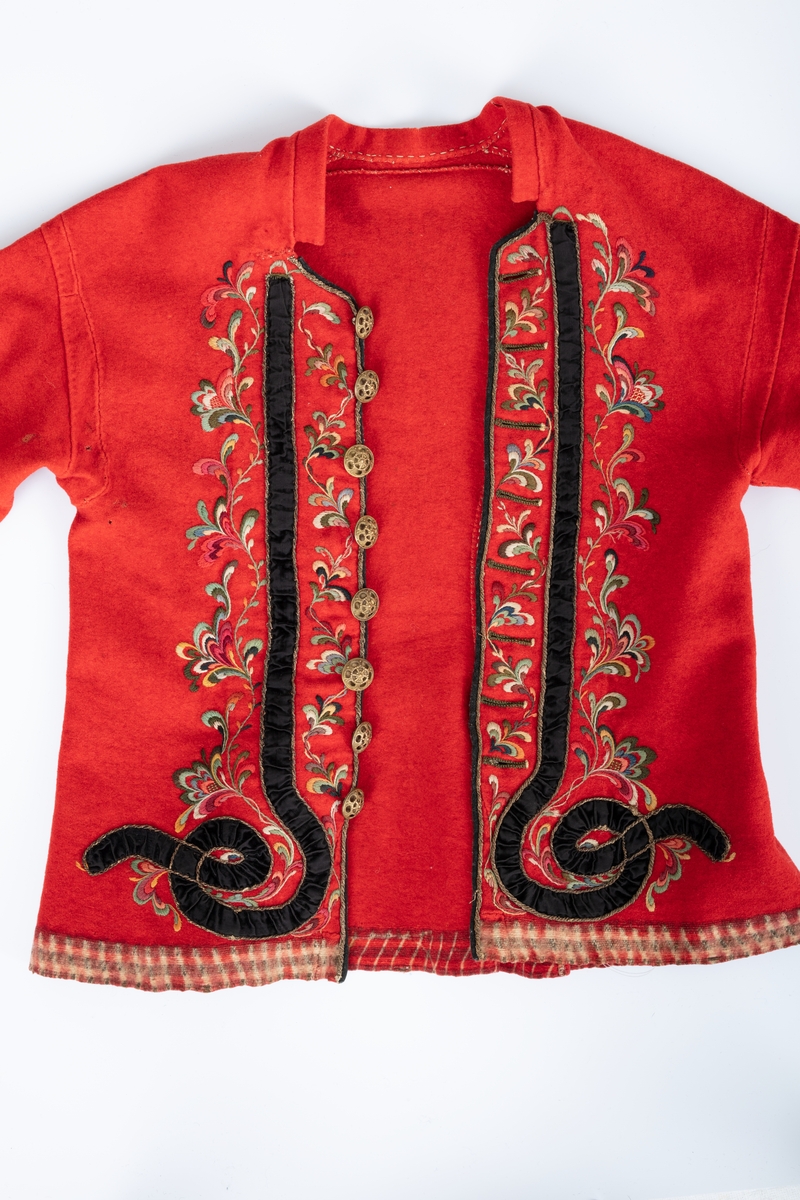 Rød trøye av klede med flerfarget ullgarnsbrodri og pynteband av svart føyel og sølvsnor.
Ermer har takkete bånd i rødt og grønt.