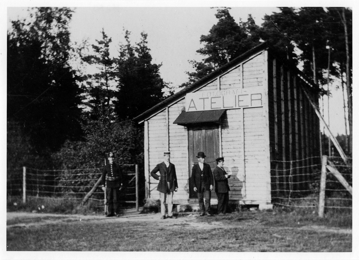 4 män i olika utstyrslar står utanför en mindre byggnad med lutande tak. Skylt ovanför dörren med text "Fotografi-Atelier". Troligen fotograf Richard Sandbergs filialateljé i Fristad hed.
