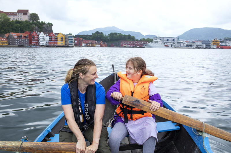 En voksen dame og en jente, begge med redningsvester, ror hver sin åre i en robåt på vannet ved Norges Fiskerimuseum