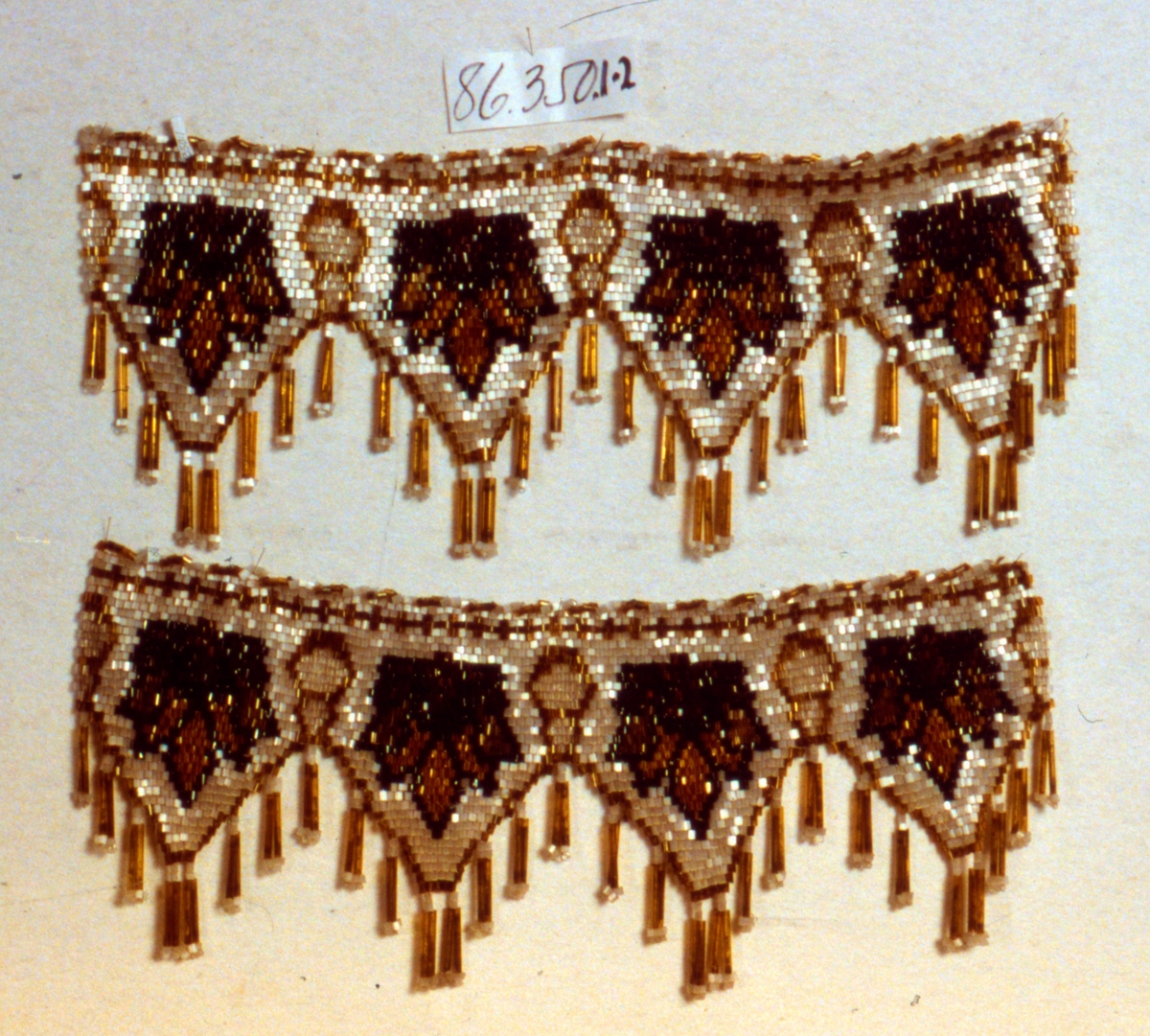 Hörnhylla med pärlfrans med ljusgrå botten med mönster i brunt och svart. Fyra uddar på varje remsa med nedhängande långa, bruna rörpärlor med små pärlor i ändarna.